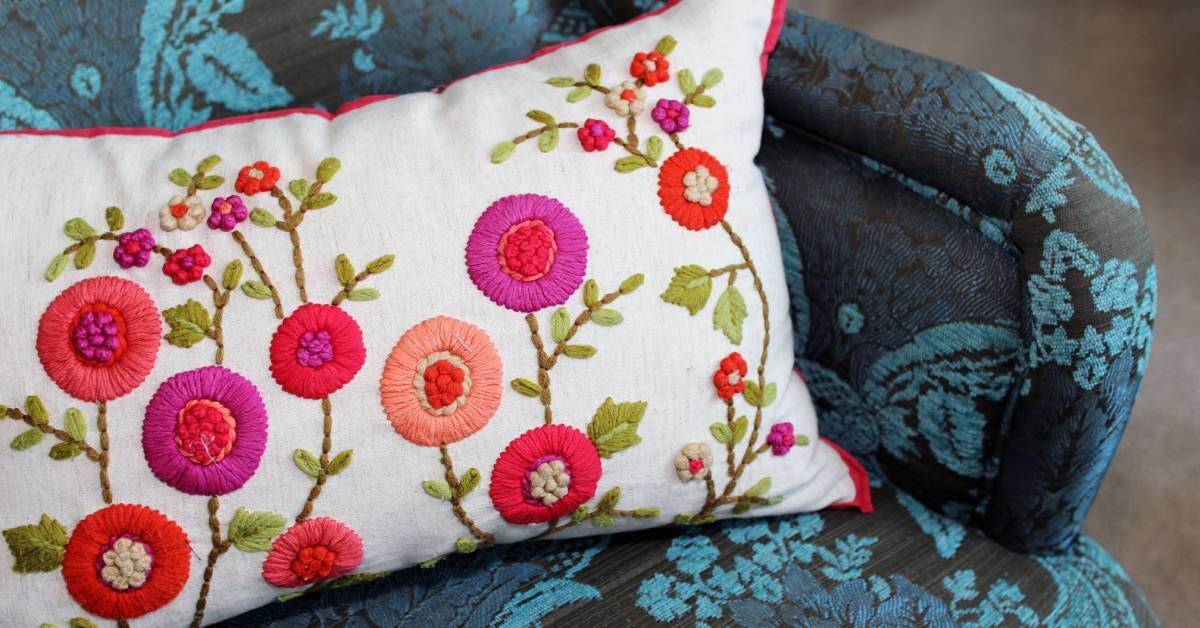 A white pillow embroidered with pink and purple flowers sitting on a black and blue embroidered vintage chair.