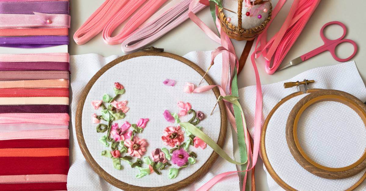 An embroidery hoop with a pink floral pattern surrounded by ribbons and embroidery tools in various shades of pink.