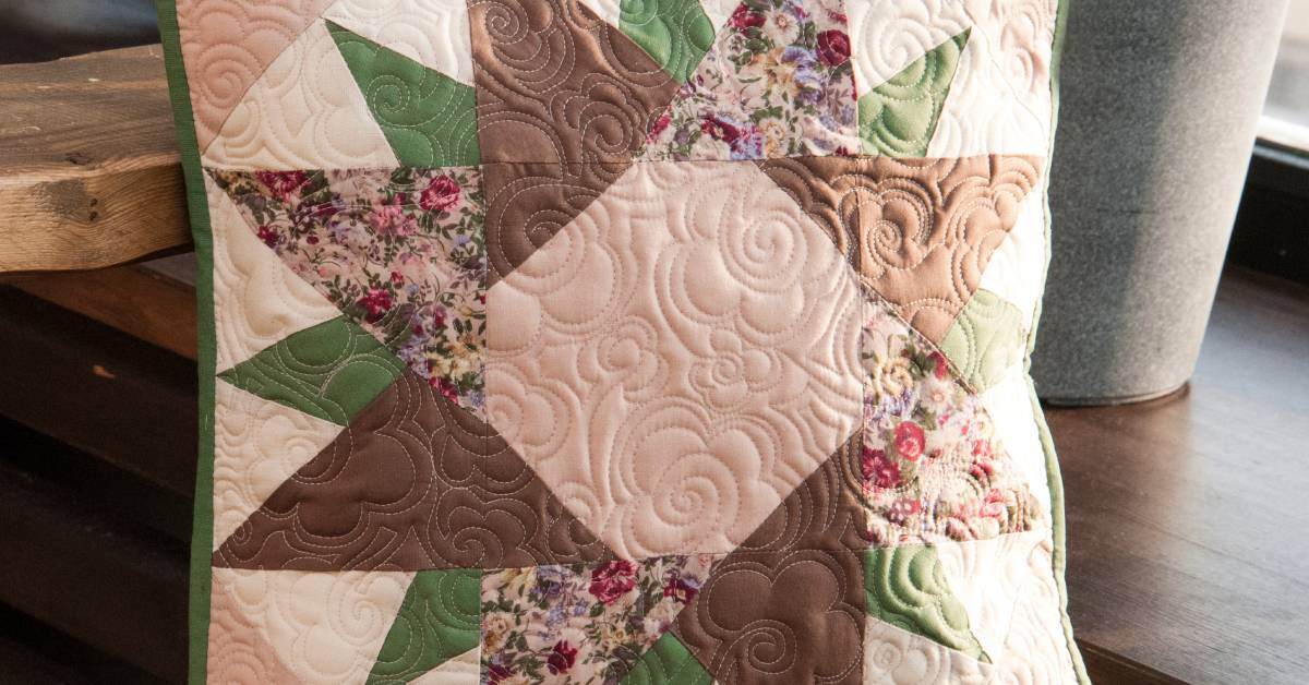 A pillow with a quilted design contains green, brown, and floral fabrics shaped to make a starburst pattern.